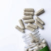 Beige herbal supplement capsules spilling from a clear plastic bottle on a white background.
